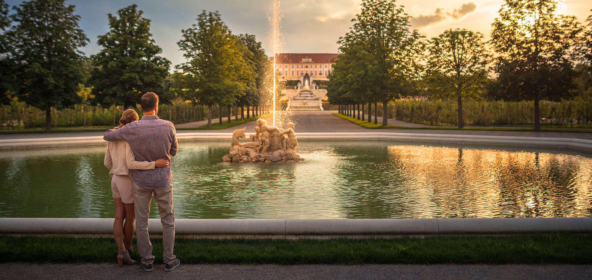 Paar steht vor dem Oktonbrunnen und blickt auf Schloss Hof während die Sonne untergeht