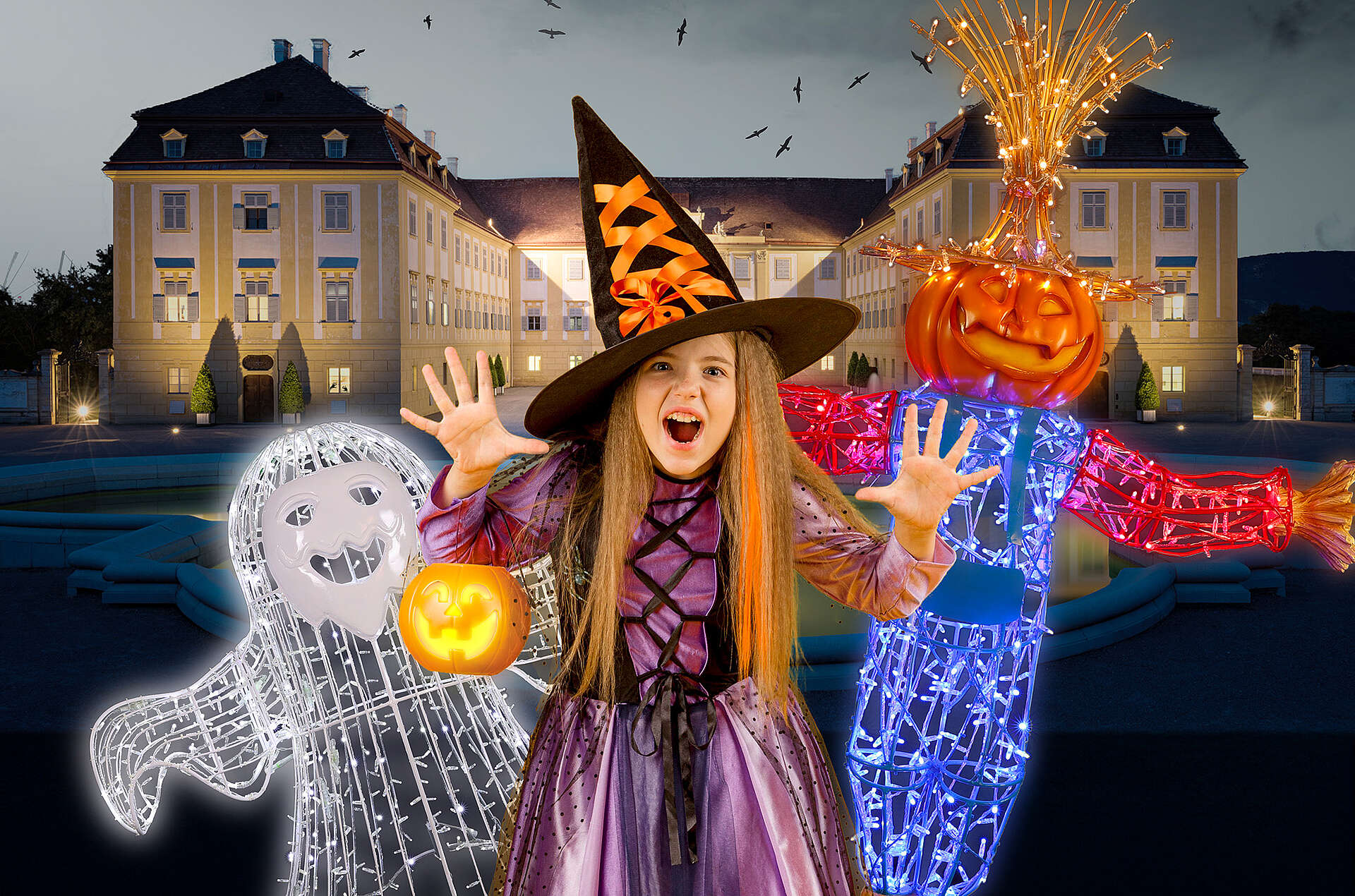 Mädchen als Hexe verkleidet, links von ihr ein Geist und rechts von ihr ein Figur mit Kürbiskopf, im Hintergrund sieht man Schloss Hof bei Nacht